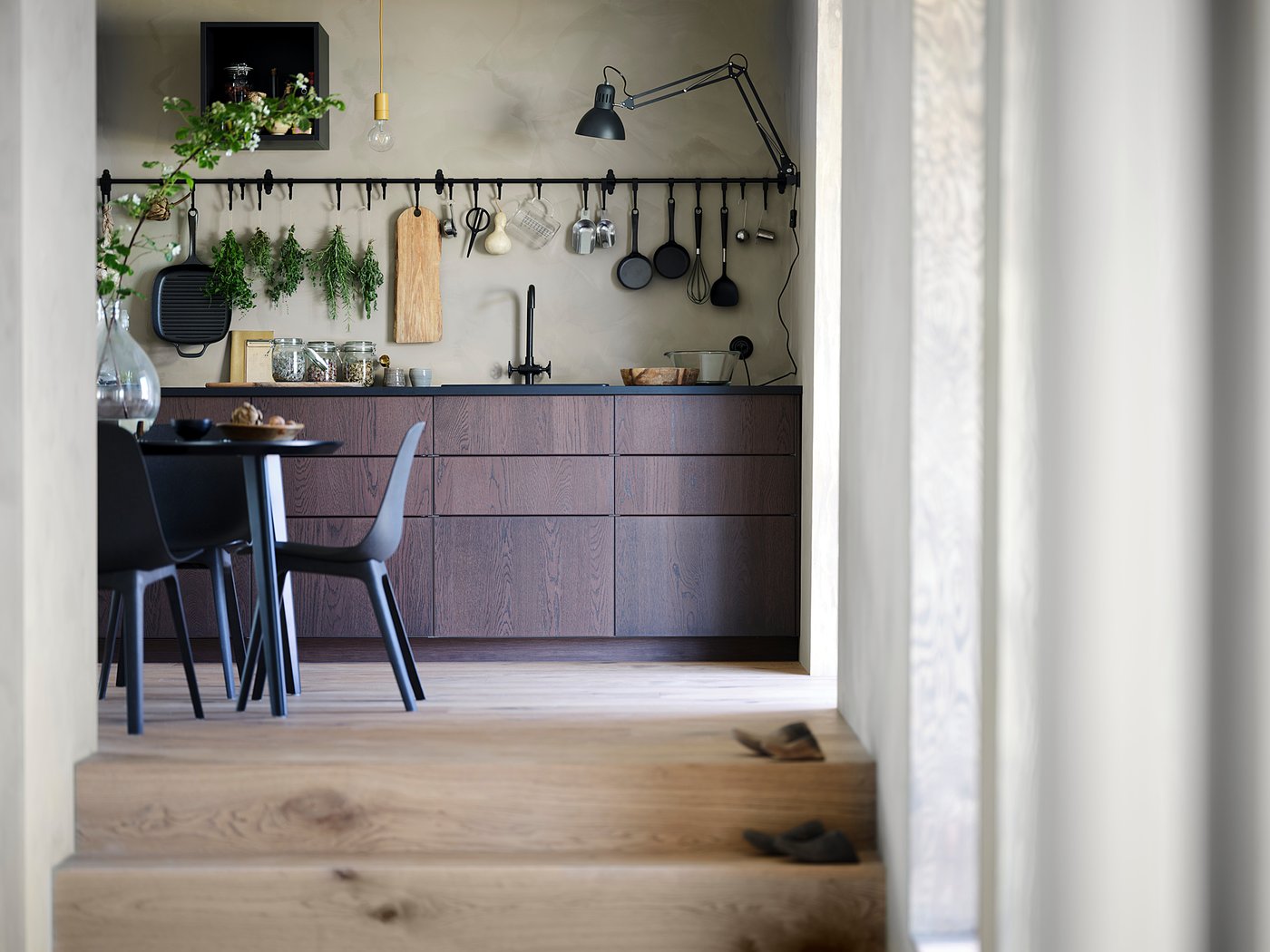 Modern kitchen with dark brown cabinets, hanging utensils, and black chairs.