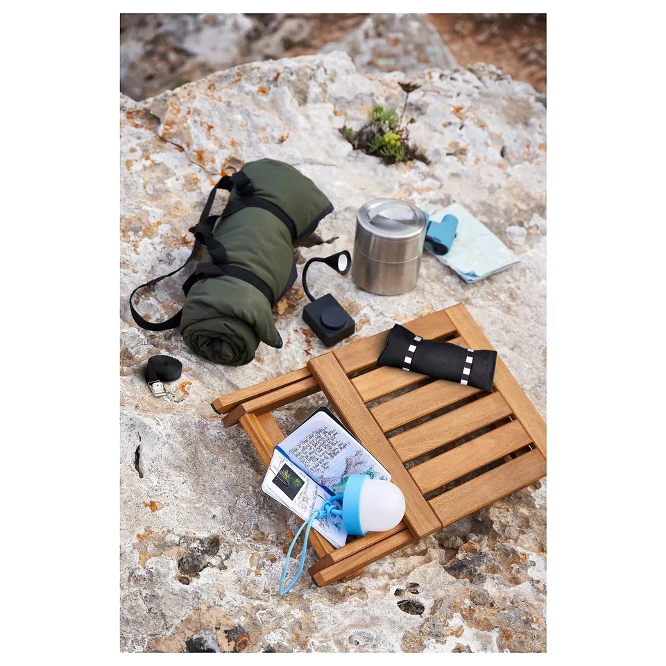 NÄMMARÖ outdoor set: folded green mat, metallic cup, map, wooden stool with blue earphones, black and white towel, and book on rocky surface.