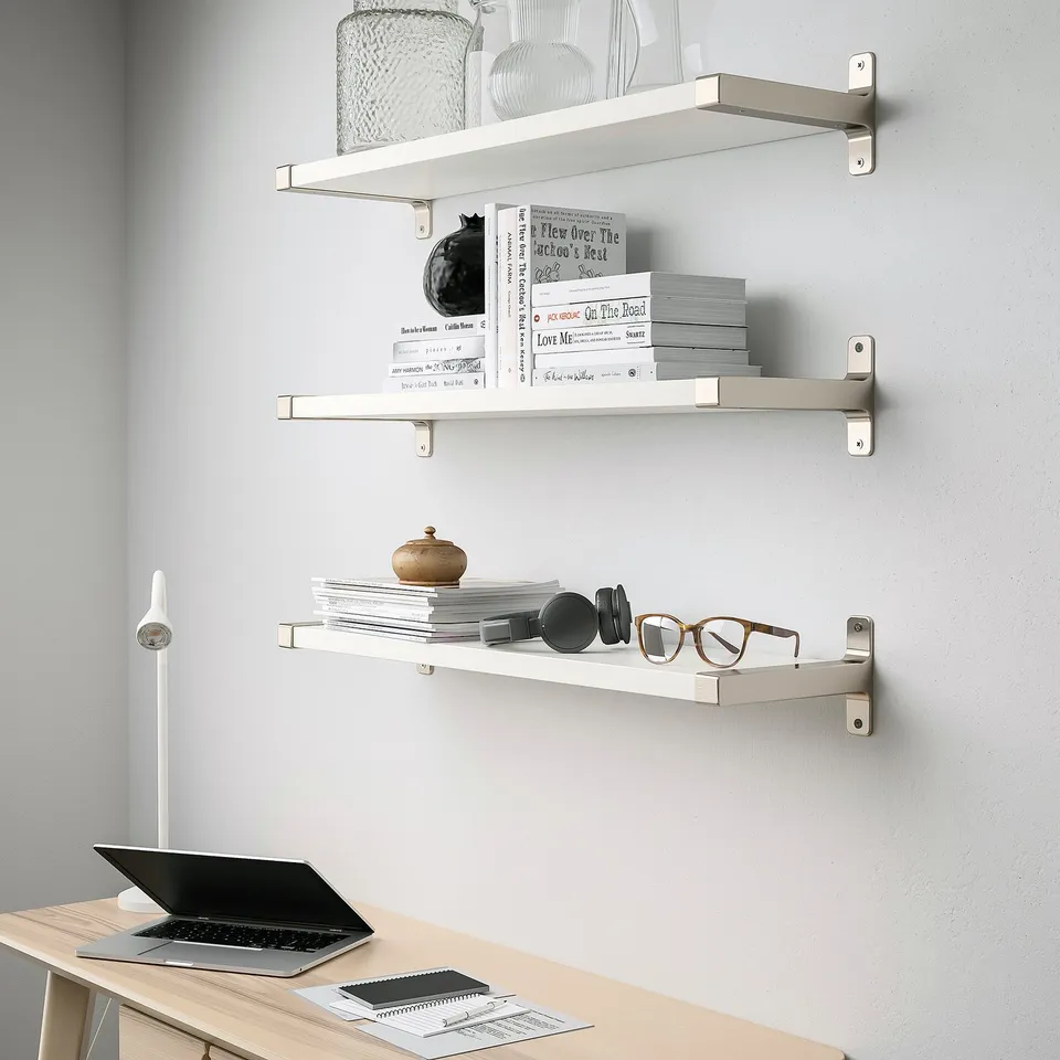 Three white wall shelves mounted above a desk. Shelves hold books, decor, and glasses. Desk has a laptop and notebook.
