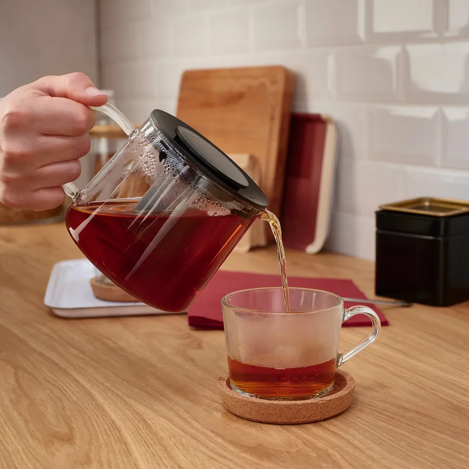 Hand pours red tea from RIKLIG glass teapot with infuser into a clear mug on a wooden table.