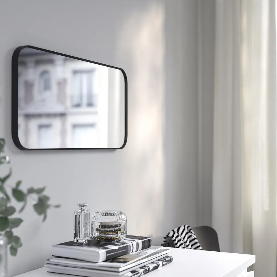 A modern mirror on a wall reflects a building, with books, a jar, and a plant below.