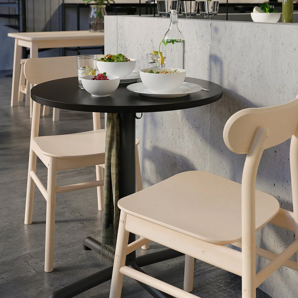 Modern café table, STENSELE, black, round, minimalist design. White chairs surround it, holding bowls of fruit and vases with flowers.