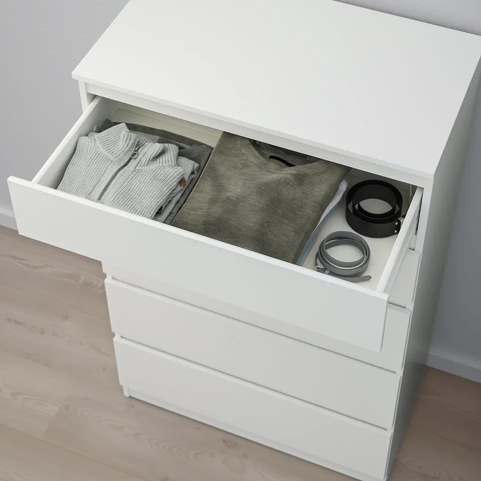 White chest of drawers with one drawer open, revealing neatly folded clothes and accessories. Safety anchoring included.
