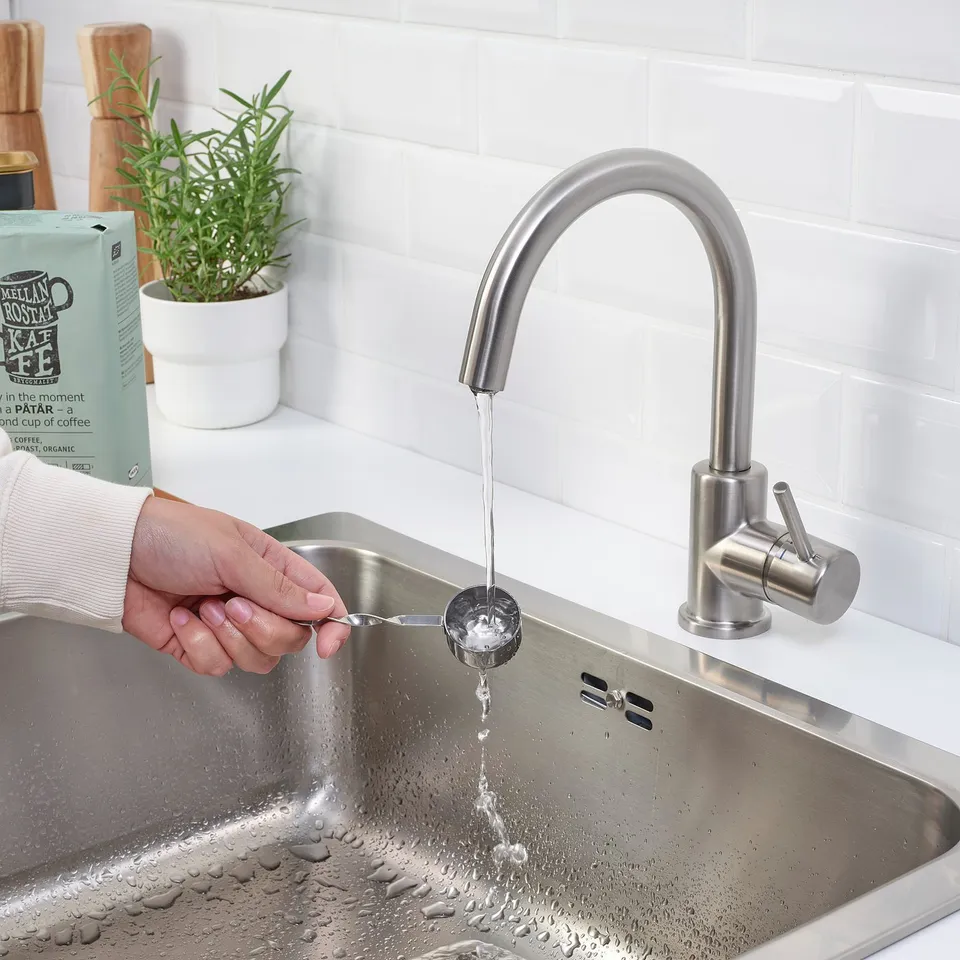 Person fills metal cup under modern chrome kitchen tap. Stainless steel sink, white tiles, and green plant visible.