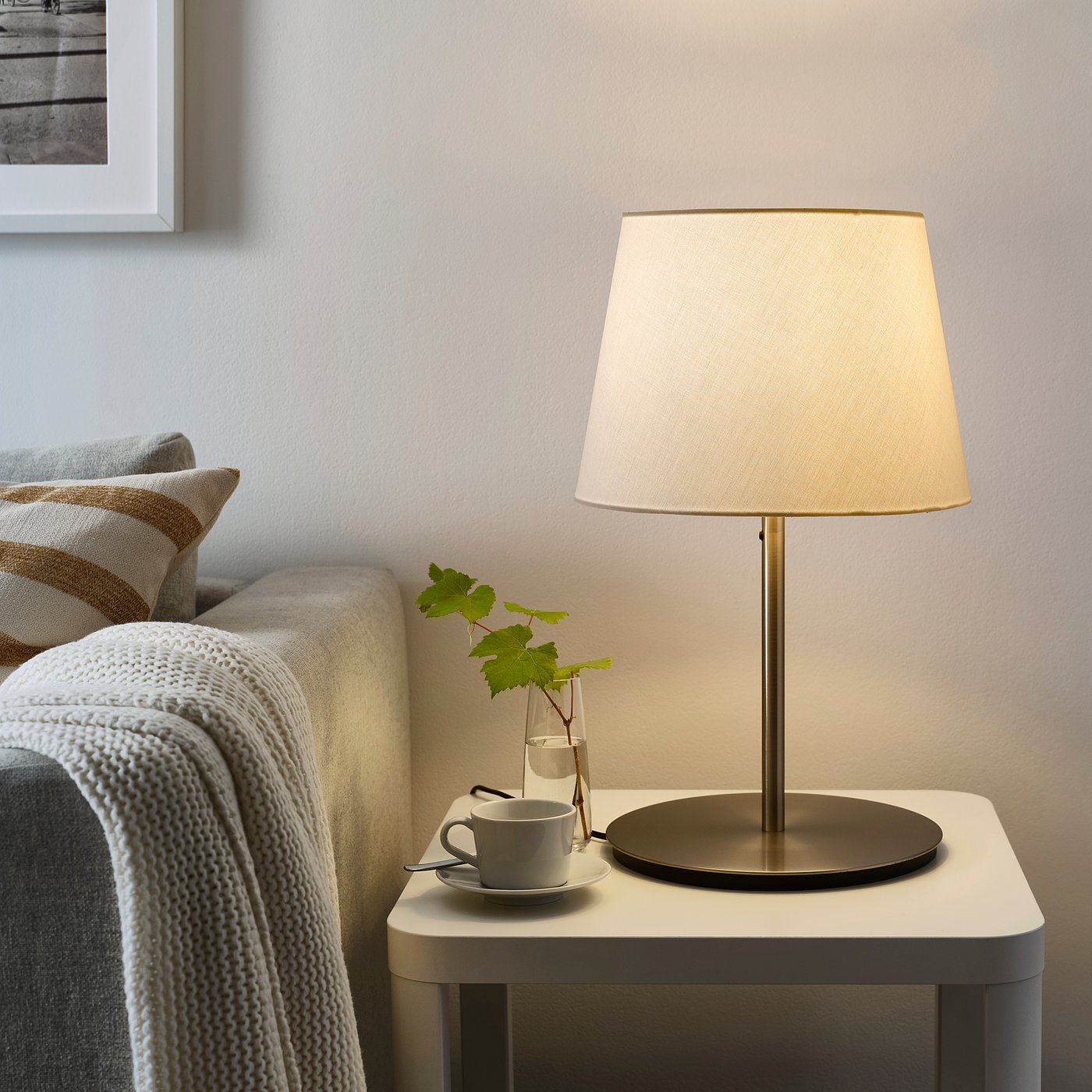 A SKOTTORP lamp with beige shade on side table, surrounded by cup, vine leaves, and cosy couch with white blanket. Soft glowing plastic shade.
