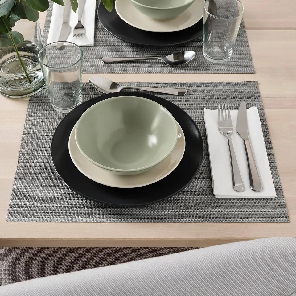 Two place settings with plain green bowls, white plates, glasses, and cutlery on woven placemats.