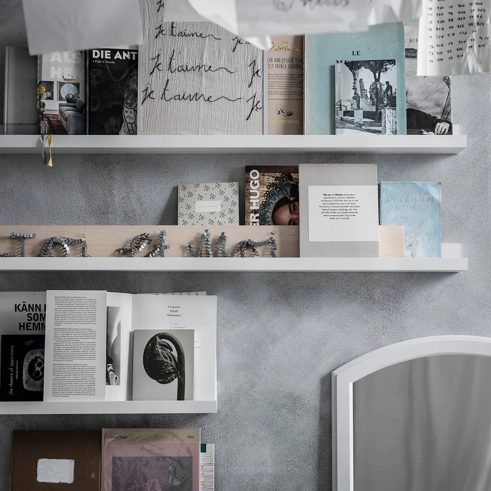 Bookshelves with books, a clock, candles, and decorative items on three white shelves against a grey wall.