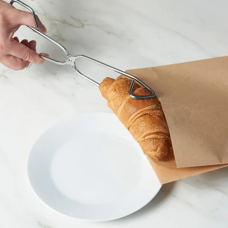 Tongs lifting a croissant from brown paper bag.