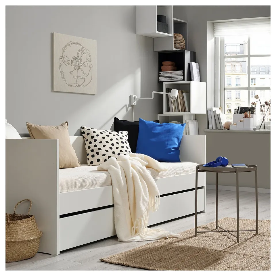 Modern white daybed with colourful pillows, blue & black, beside a metal table. Cosy blanket and minimalist decor complete inviting space.