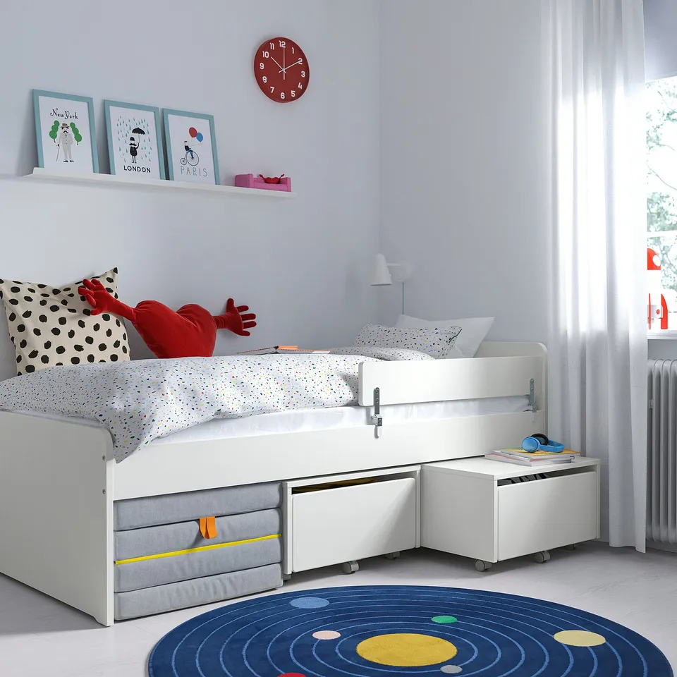 Childs bedroom feature SLÄKT white bed with guard rail, dot pillow, red doll, under-bed storage, blue rug, wall art, clock, lamp, and radiator.
