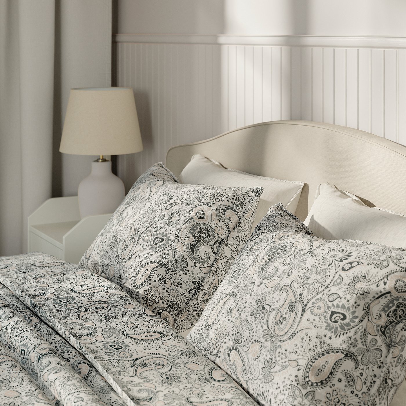 Bed with upholstered headboard, intricate paisley print bedding, and a bedside lamp on a white bedside table.