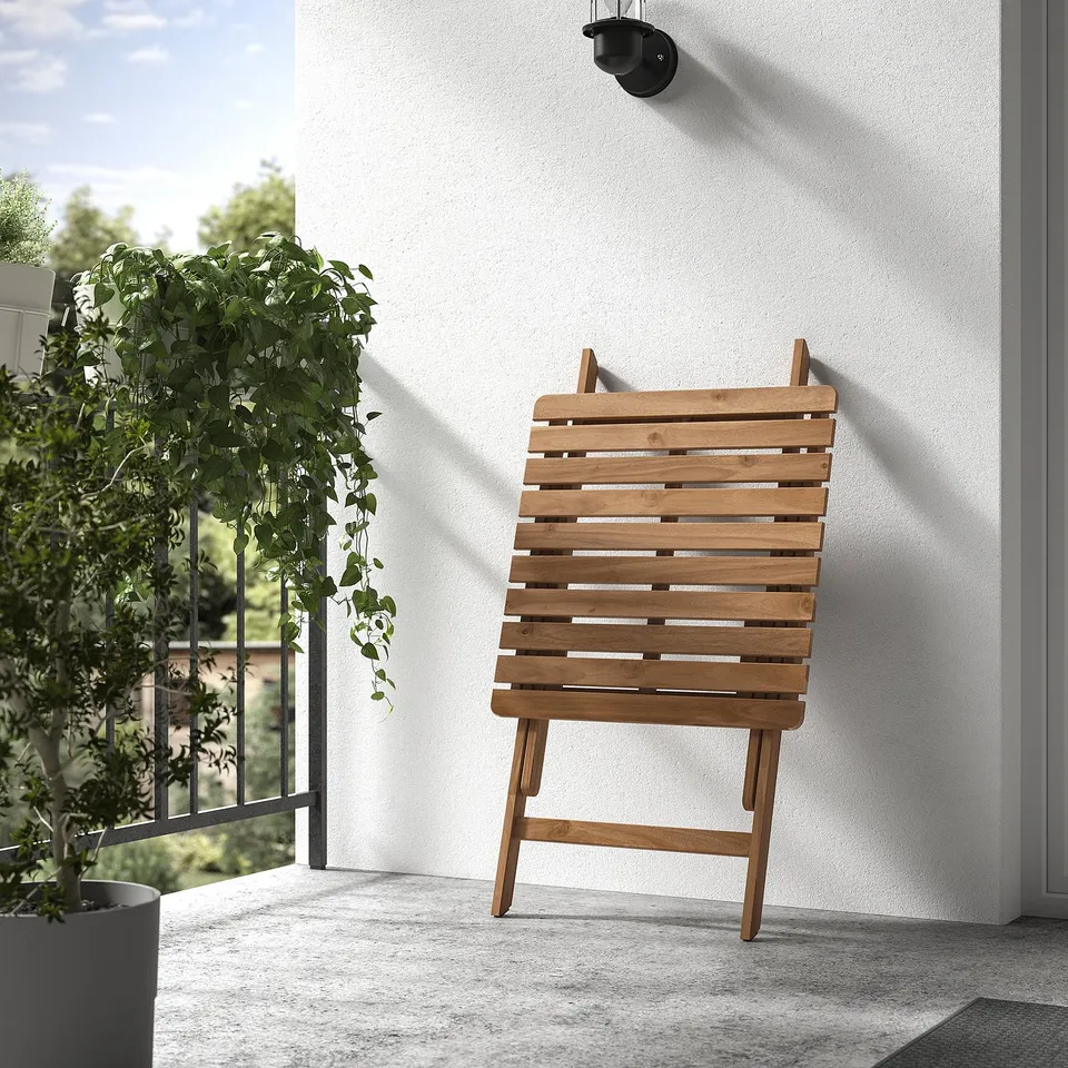 Wooden folding chair leaned against white wall near green plant, perfect for outdoor use.