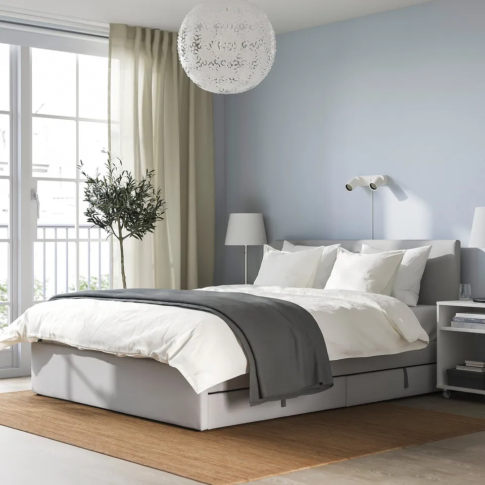 Modern grey GLADSTAD bed dominates room, white linens, wooden floor, large window with tree, round light.