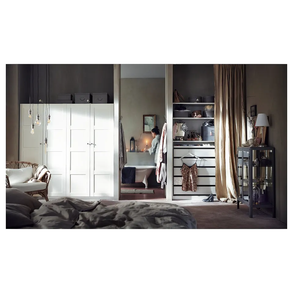 Modern bedroom with white wardrobes, pendant lights, beige curtains. Unmade bed on left, freestanding bathtub centre with person beside it.