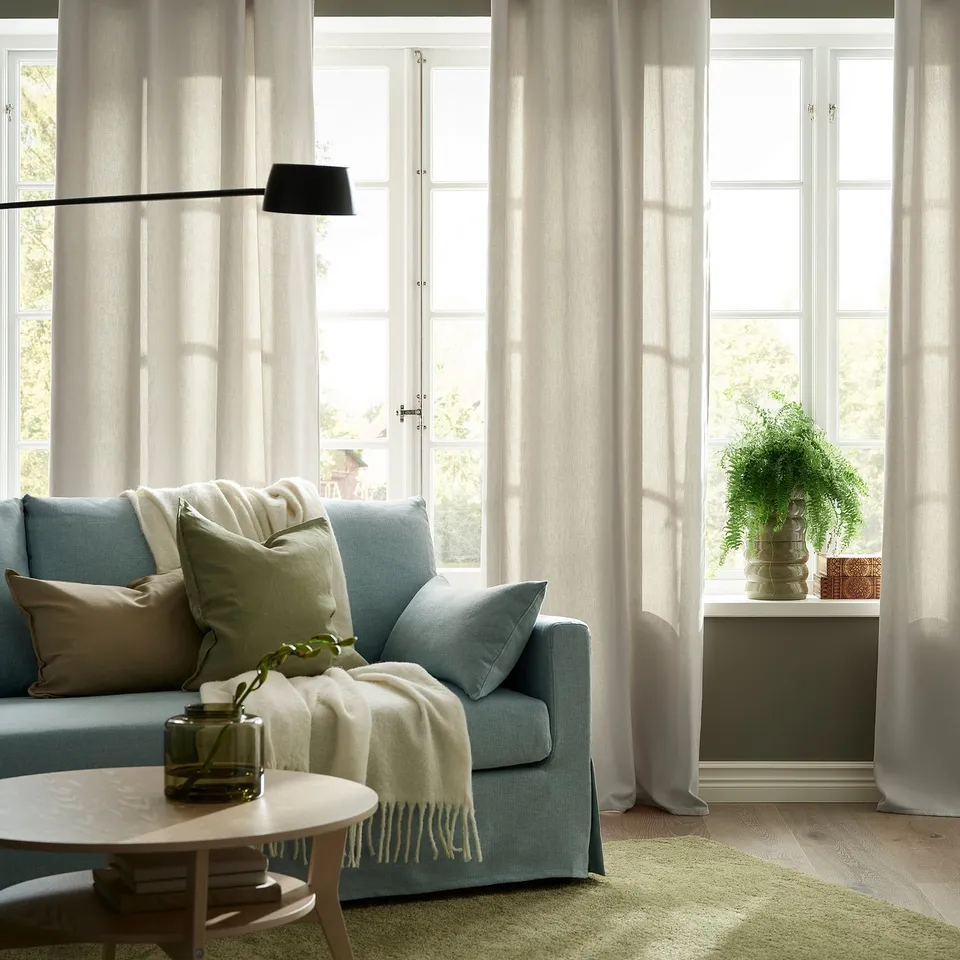Light-filled room with light blue couch, beige GINSTMOTT curtains, and plant. Curtains softly philtre light, providing privacy.