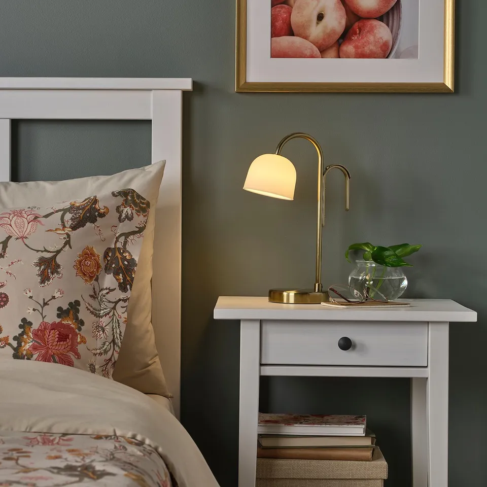 Small bedside lamp, TVÅMASTAD, brass-colored, frosted glass shade, atop tiny white table; floral pillow, grey wall, books, and vase nearby.