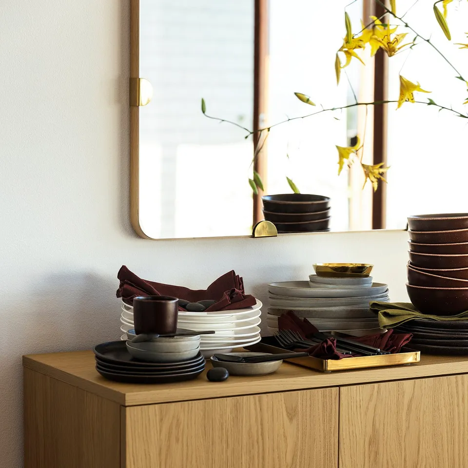 Wooden cupboard with plates, cutlery, and napkins; mirror above. Black & white dishes, maroon cloths, fern in vase beside bowls.