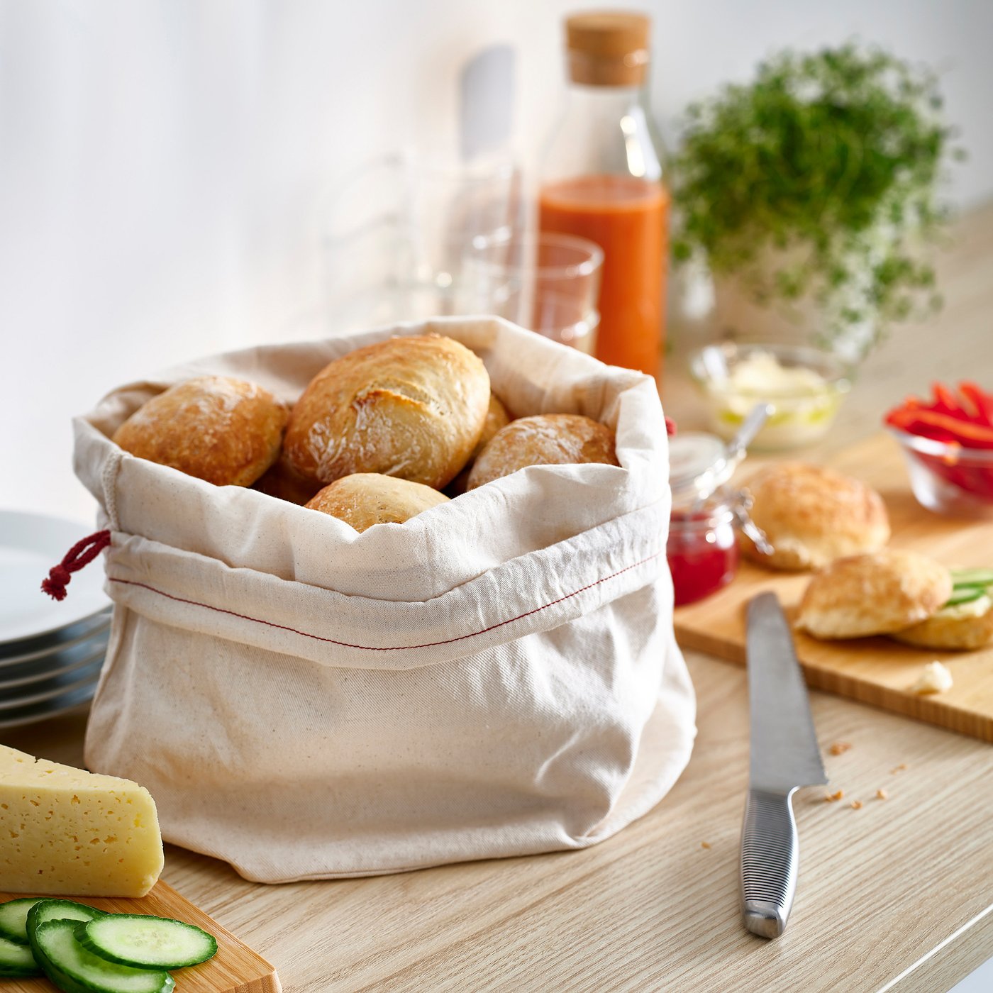 White cloth bread bag with rolls, on wooden table, next to knife, cheeses, veggies, and condiments.