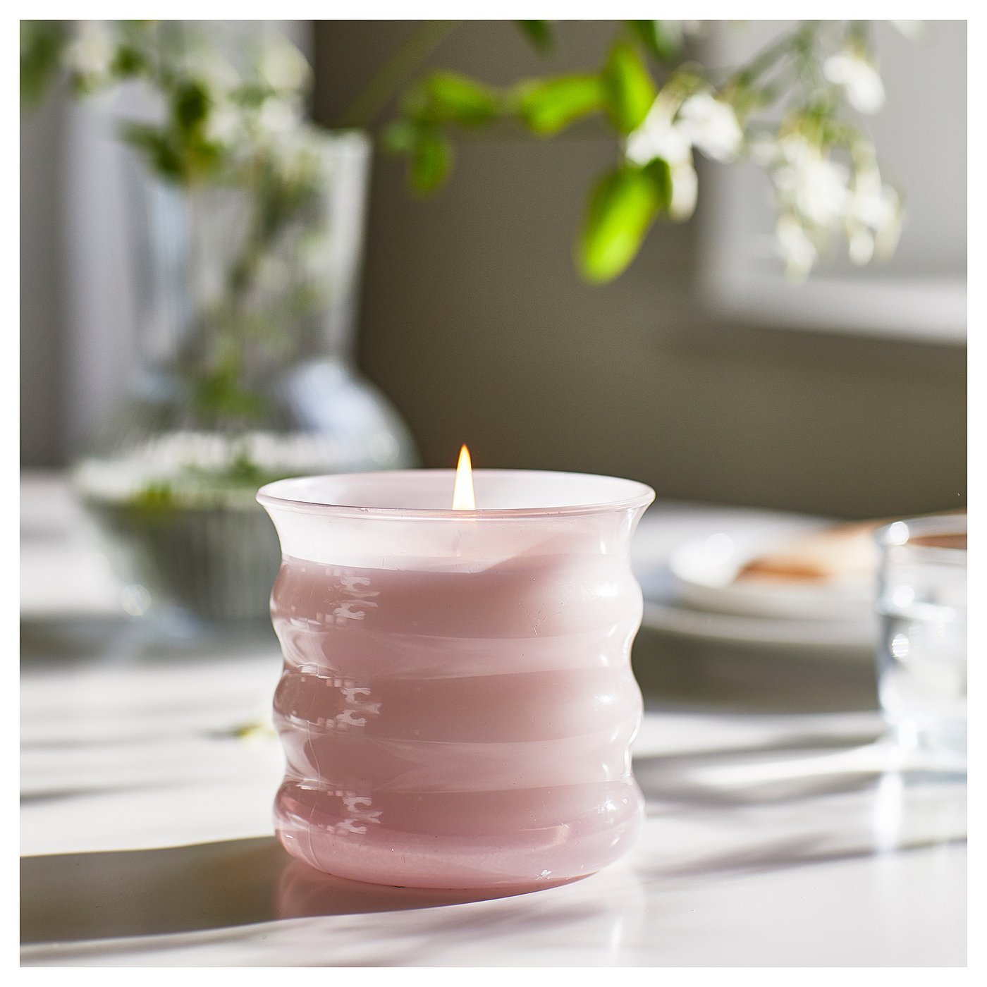 Pink frosted glass candle in ribbed design, lit, on a dining table with greenery backdrop.