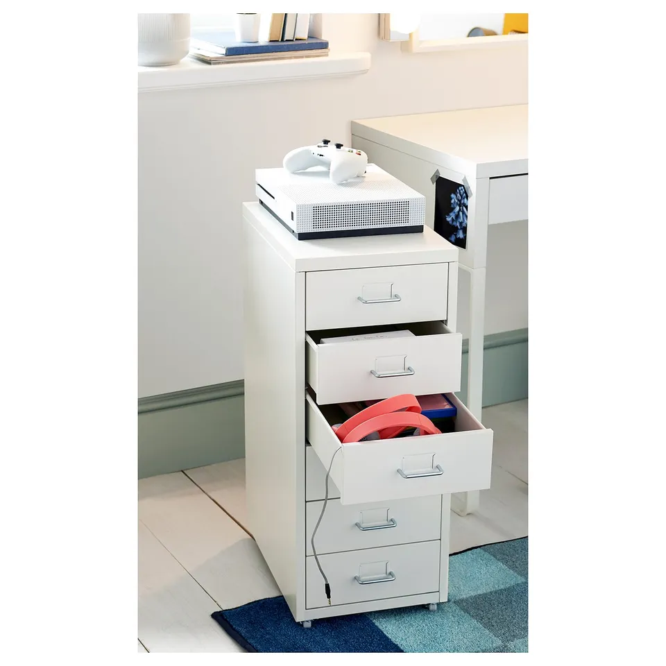 A white HELMER rolling file cabinet with drawers, used to store gaming accessories, including a console and controllers, in a clean, organised room.