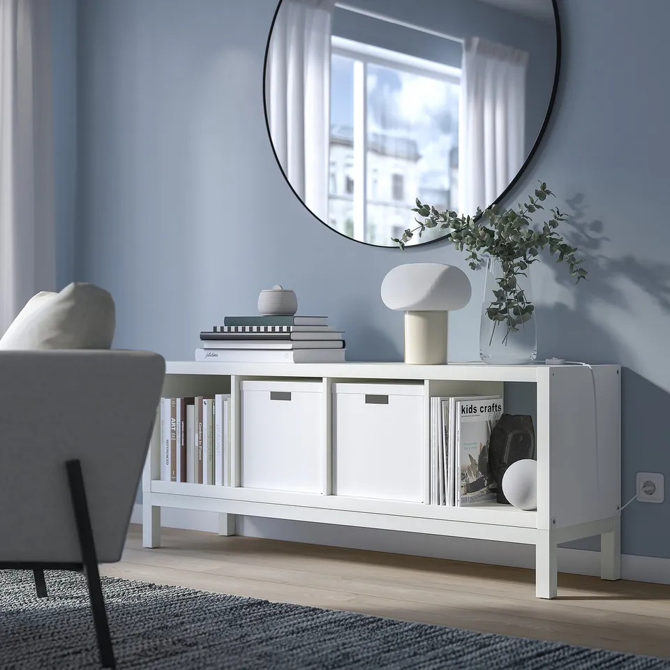 KALLAX shelf with books, decor, and lamp against blue wall, mirror reflecting window, grey chair, striped rug.