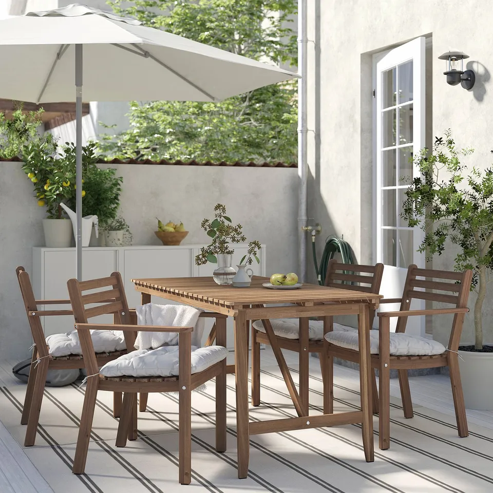 Outdoor dining setup with brown acacia table and four matching chairs, cushioned in white, shaded by a large umbrella, creating a cosy patio ambiance.