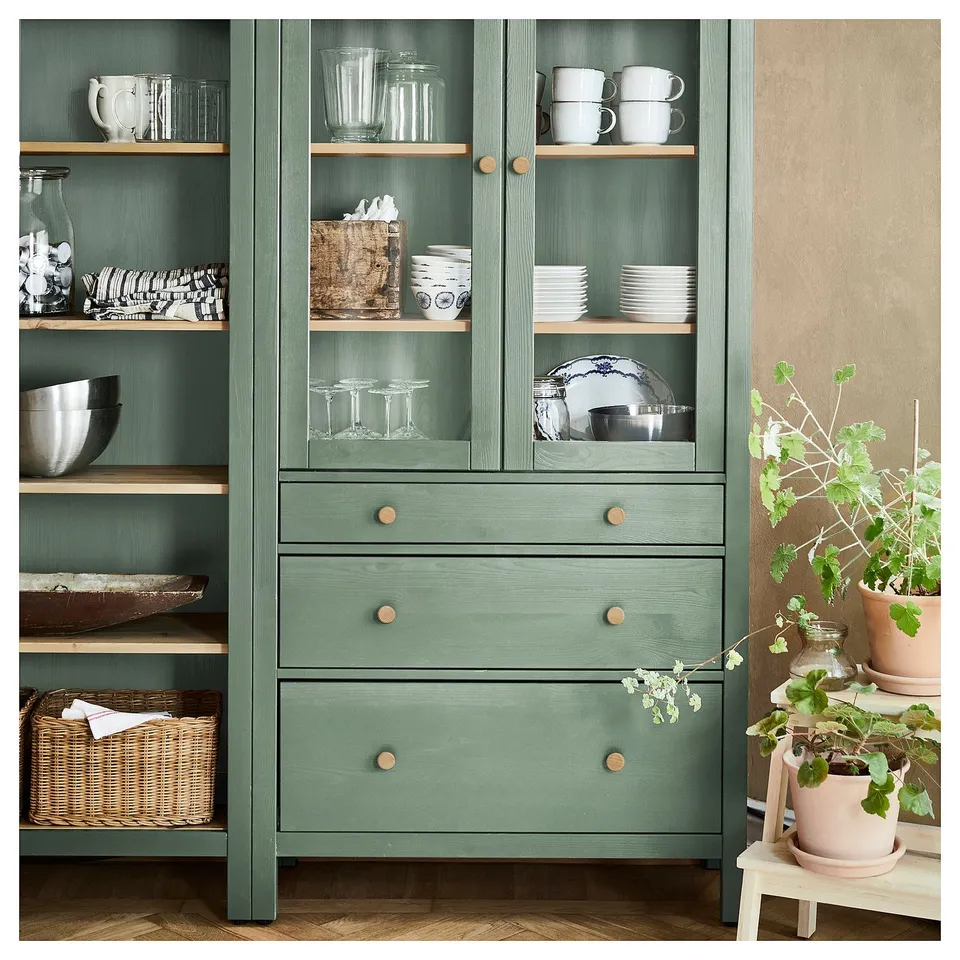 HEMNES green wooden storage unit with glass doors, shelves, and drawers, displaying dishes, cups, and plants.