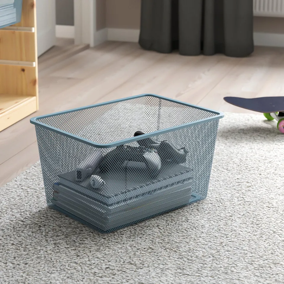Mesh storage bin on carpeted floor, containing several items, designed for easy access and organisation. Ideal for indoor use with TROFAST frames.