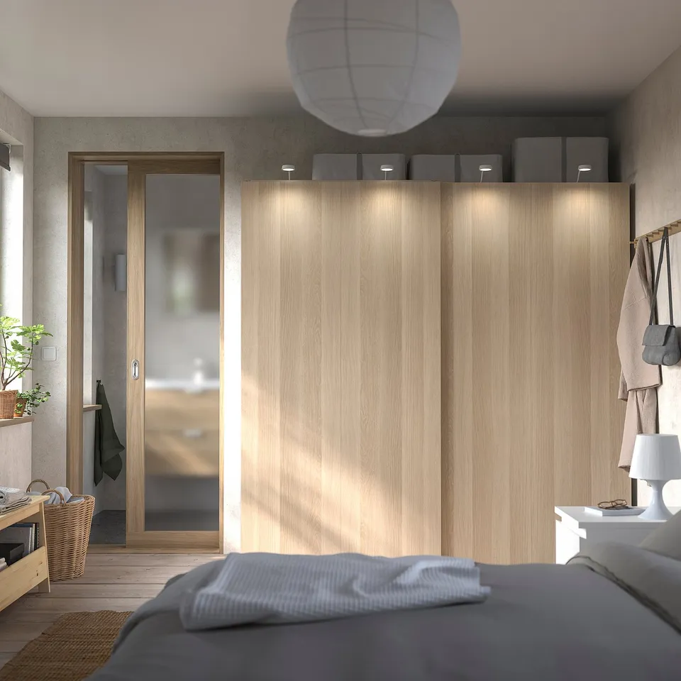 Bedroom with light wood closet, sliding door, grey bed linen, hanging sweaters, and white lamp.