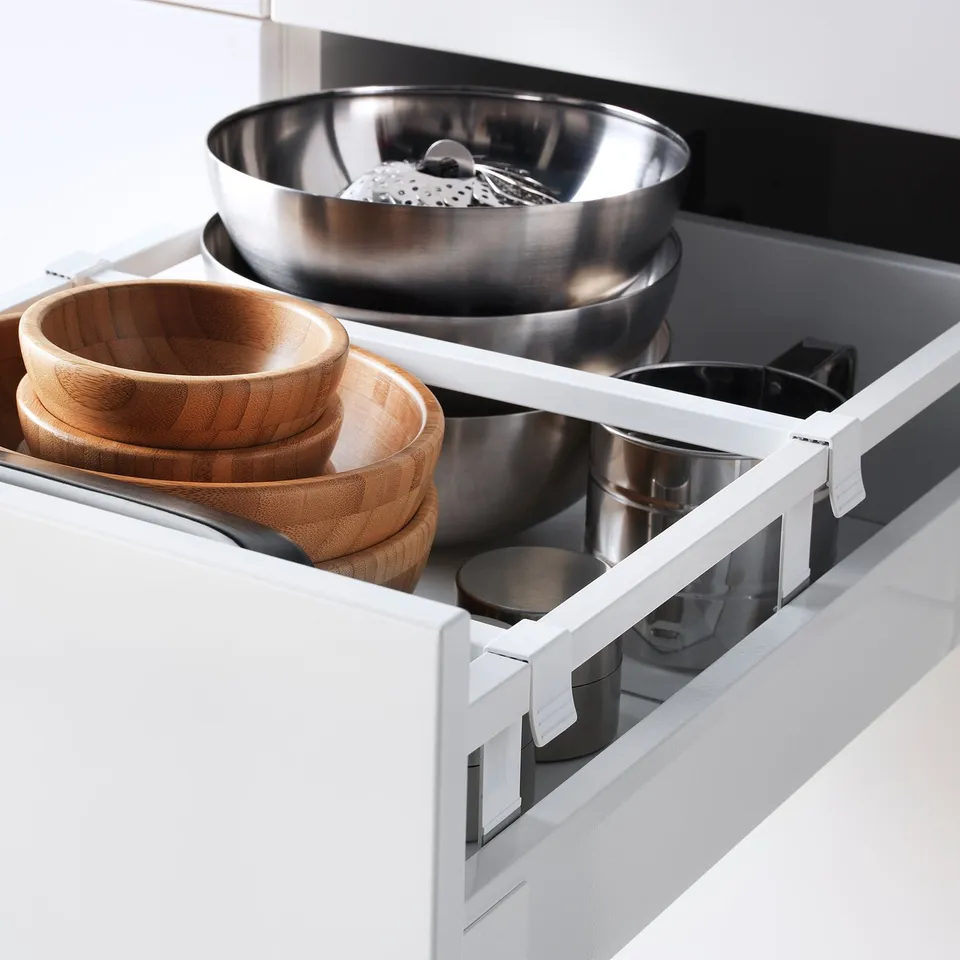 White kitchen drawer extended, showing stainless steel bowls, wooden bowls, and kitchen tools. Part of METOD system; self-closing mechanism visible.