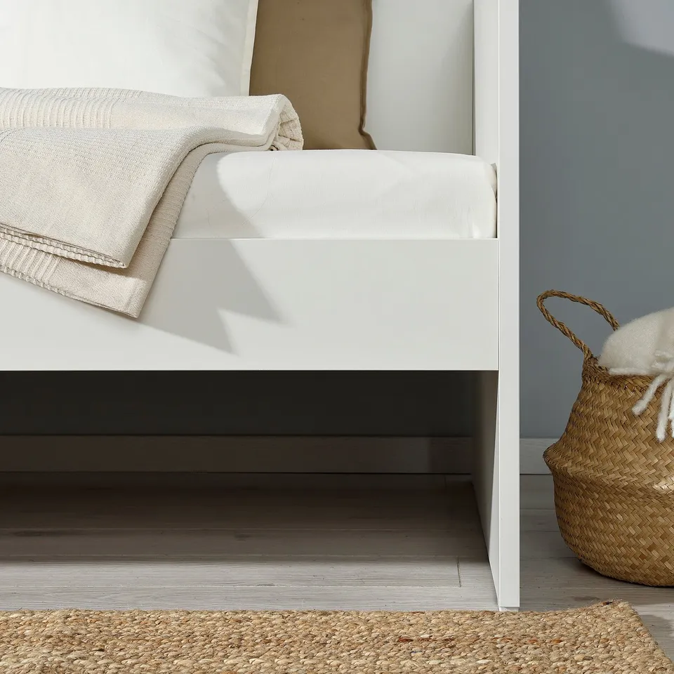 A neatly made bed with white linens against a light-colored wall. A woven basket with white items is on the floor beside it.
