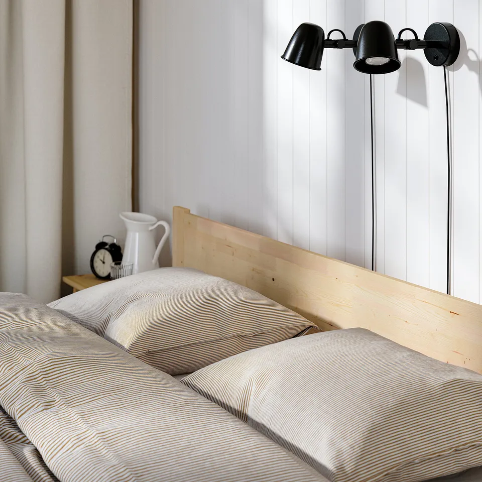 A simple, scandinavian-style wooden bed frame with adjustable wall-mounted reading lamps beside it.