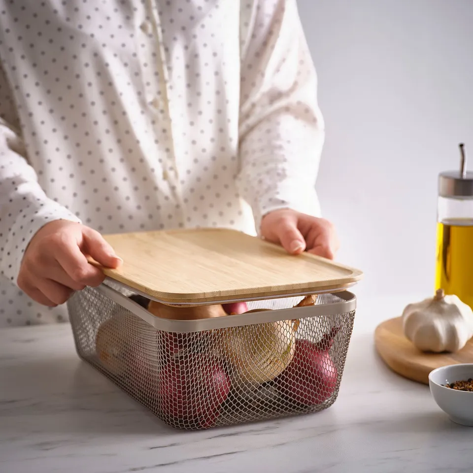 Person places wooden lid on mesh food storage basket on counter. Basket holds fruits, near oil bottle and garlic.