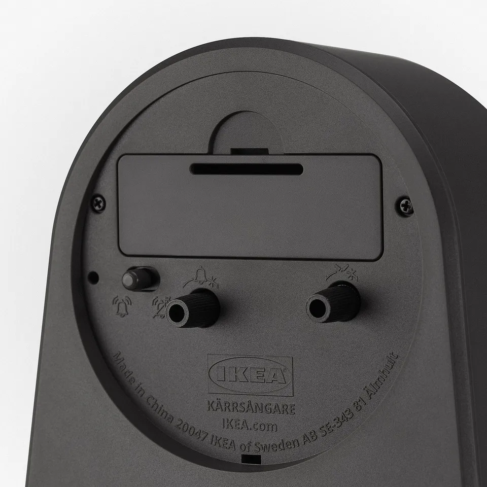 A sleek black alarm clock with a smooth finish, featuring a large button, two knobs, and a slot at the top, with text and logos on the bottom.
