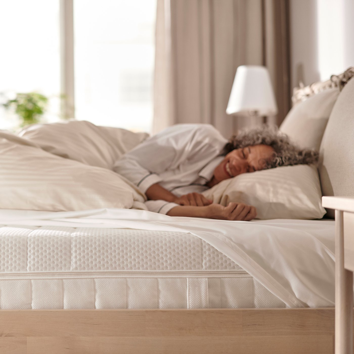 Peaceful sleeper on beige mattress in bright room with lamp & plant.