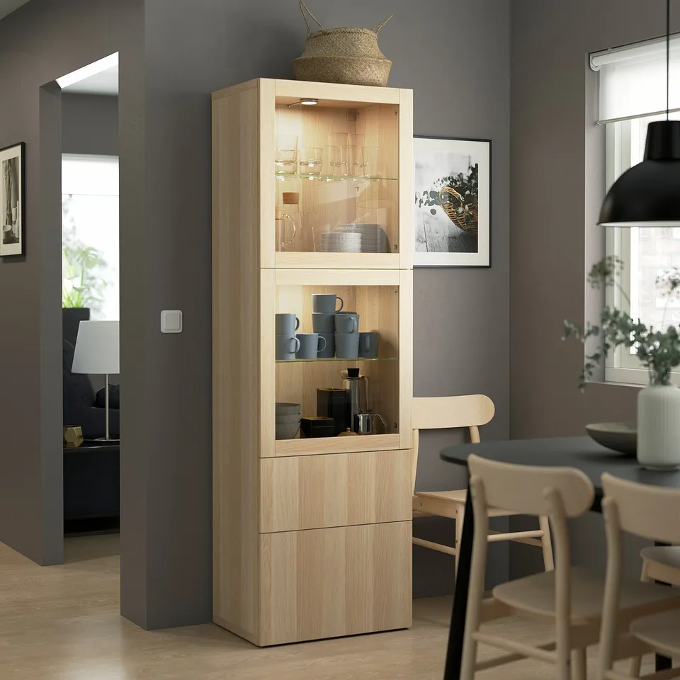 Modern, light wooden BESTÅ cabinet with glass doors, holding mugs, showing in a stylish dining area.