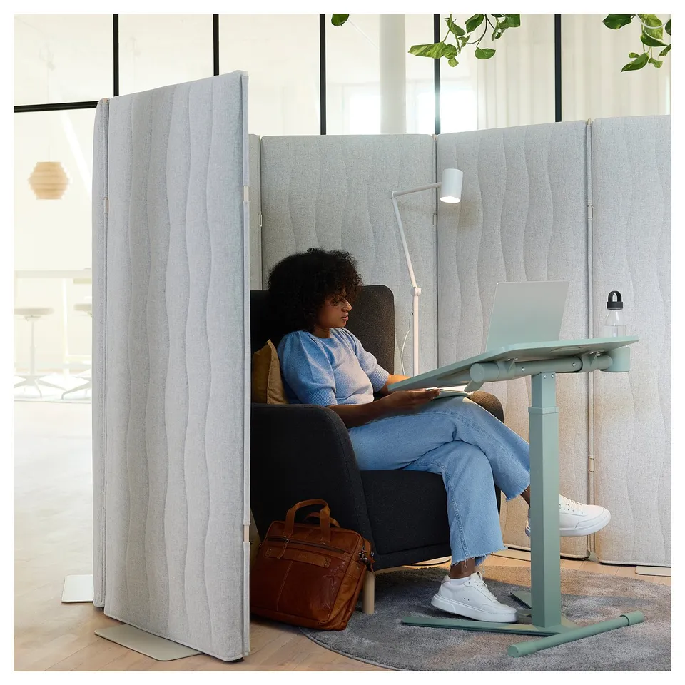 A person uses a laptop in a private work zone created by a grey fabric acoustic screen. A light blue desk, chair, and brown bag complete the setup.