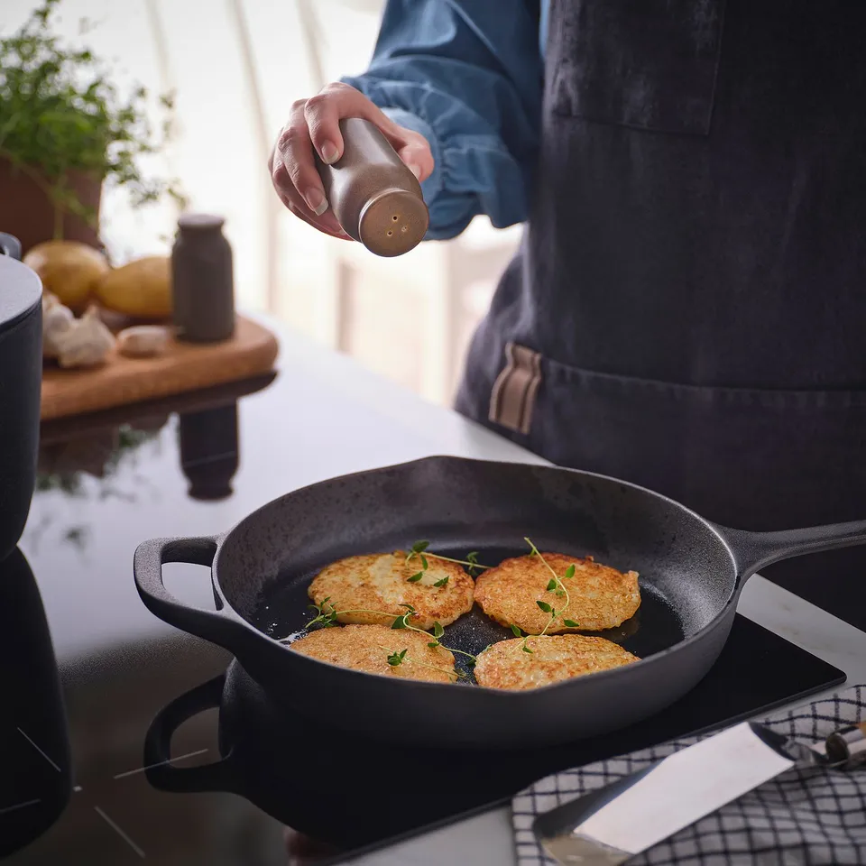 A person seasoning food in a skillet with a salt shaker from the BRUGDHAJ series.