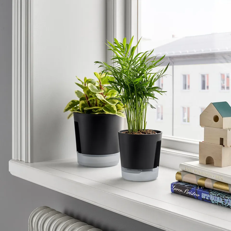 Two black VÅRDTRÄD self-watering plant pots on shelf with plants, books, wooden decor near window