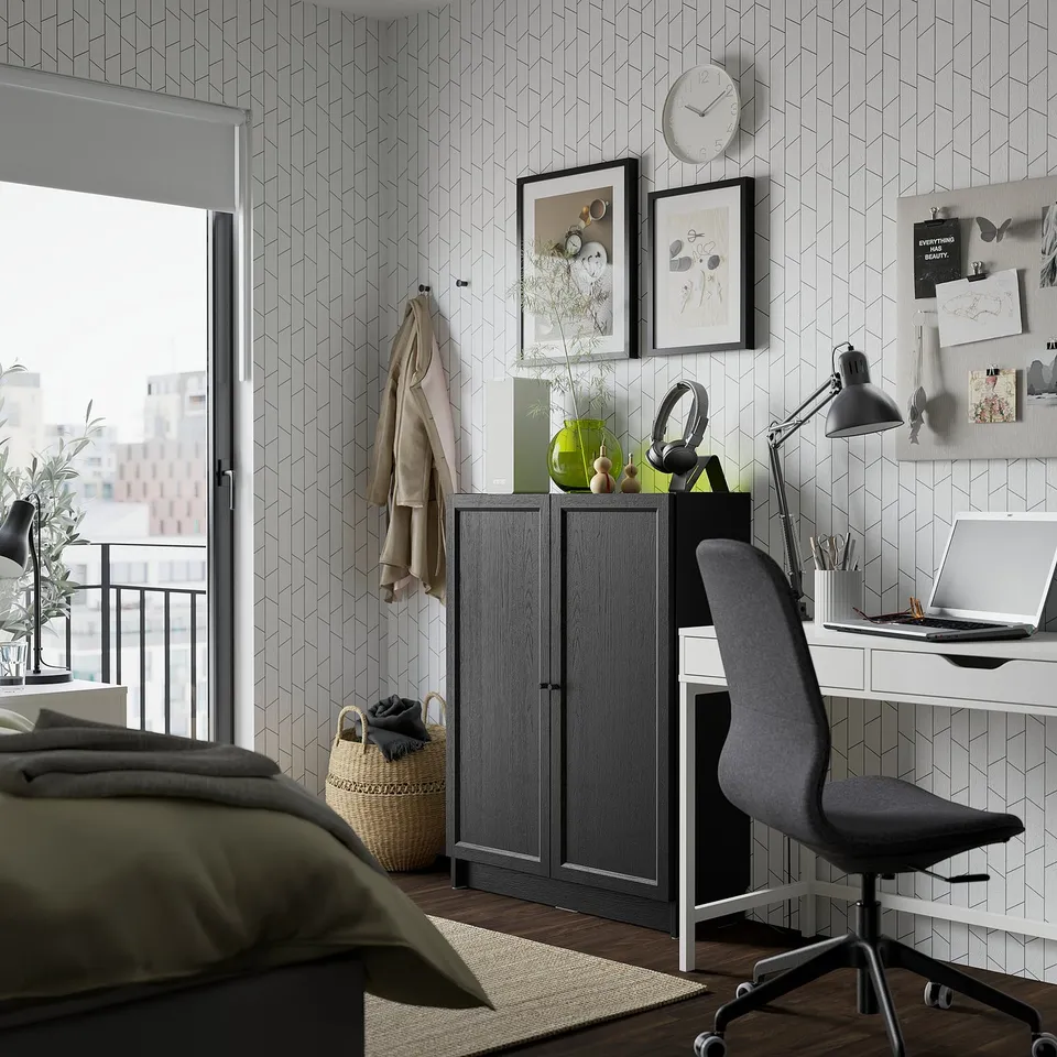 A modern bedroom BILLY bookcase in black-brown with adjustable shelves. It stands between a white desk and a bed, holding decorative items.