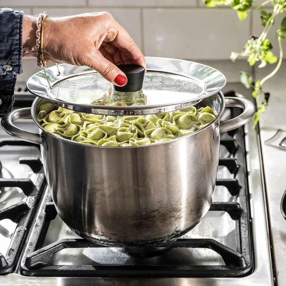 Cooking scene with KLOCKREN lid, clear glass, silver rim, on stove. Cheese and herbs nearby.