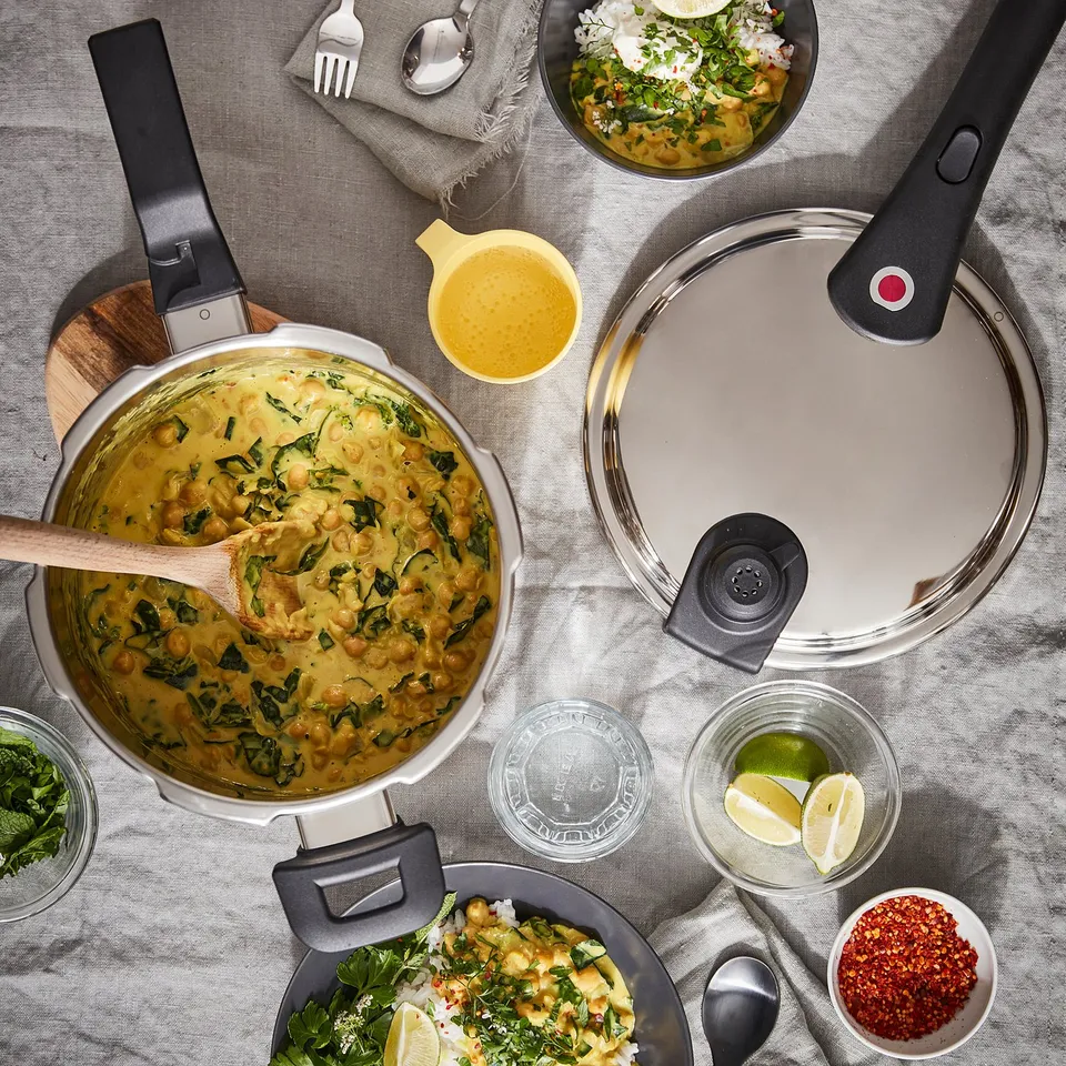 Cooking scene with IKEA 365+ pressure cooker, yellow sauce, limes, herbs, and served dishes on grey cloth.