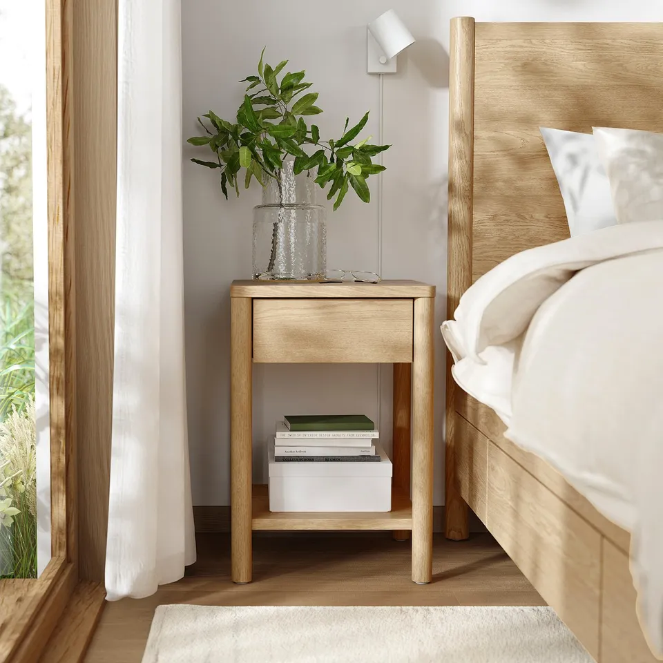 Small wooden bedside table, light colour, between bed & window. Holds vase with green plants, books, & box.