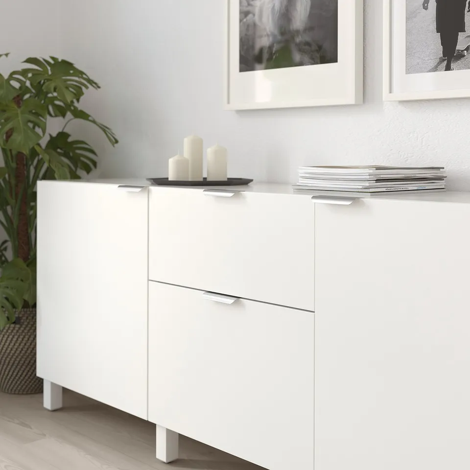White cabinet with modern handles, showcasing clean lines. Three candles on top, stacks of magazines beside, and a plant nearby.