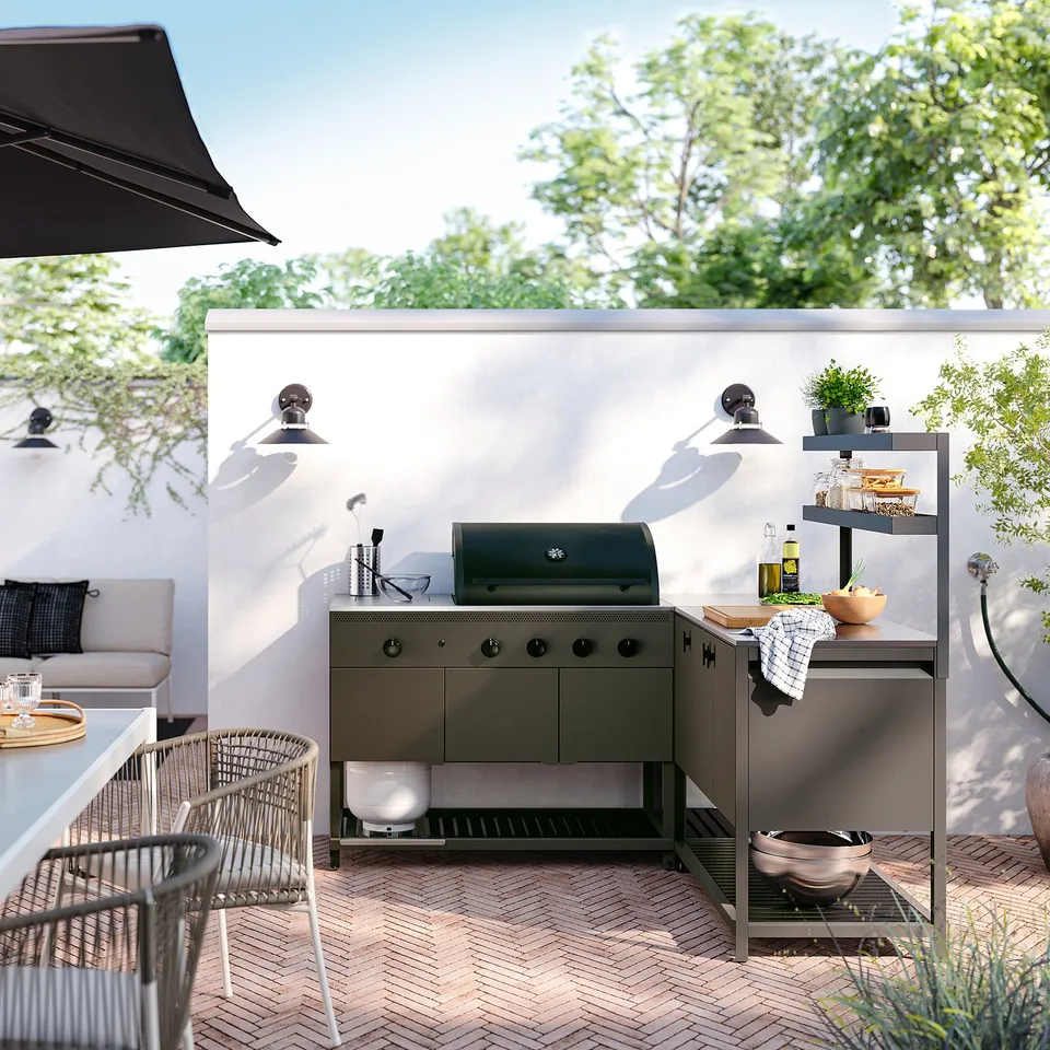 Modern outdoor kitchen with grey metal cabinets, grill, and shelves. Surrounded by patio furniture and greenery.