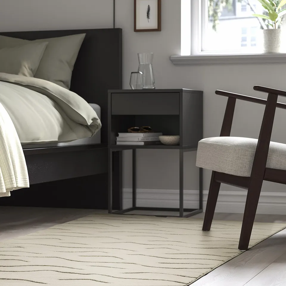 Modern bedroom with black bed, small black side table, white chair, and beige VÄXELFEL rug.