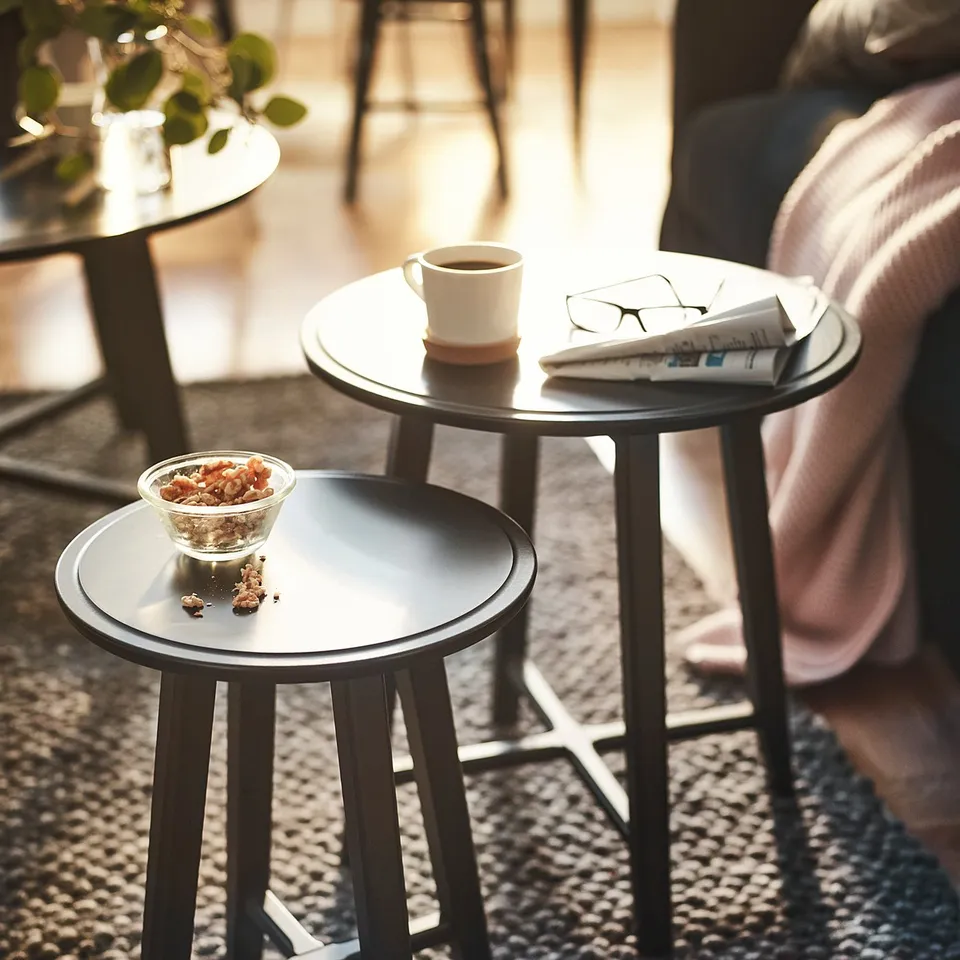 Black round nesting tables, one with a coffee cup, magazine, and glasses, the other with a bowl of snacks. Modern, sleek design for small spaces.