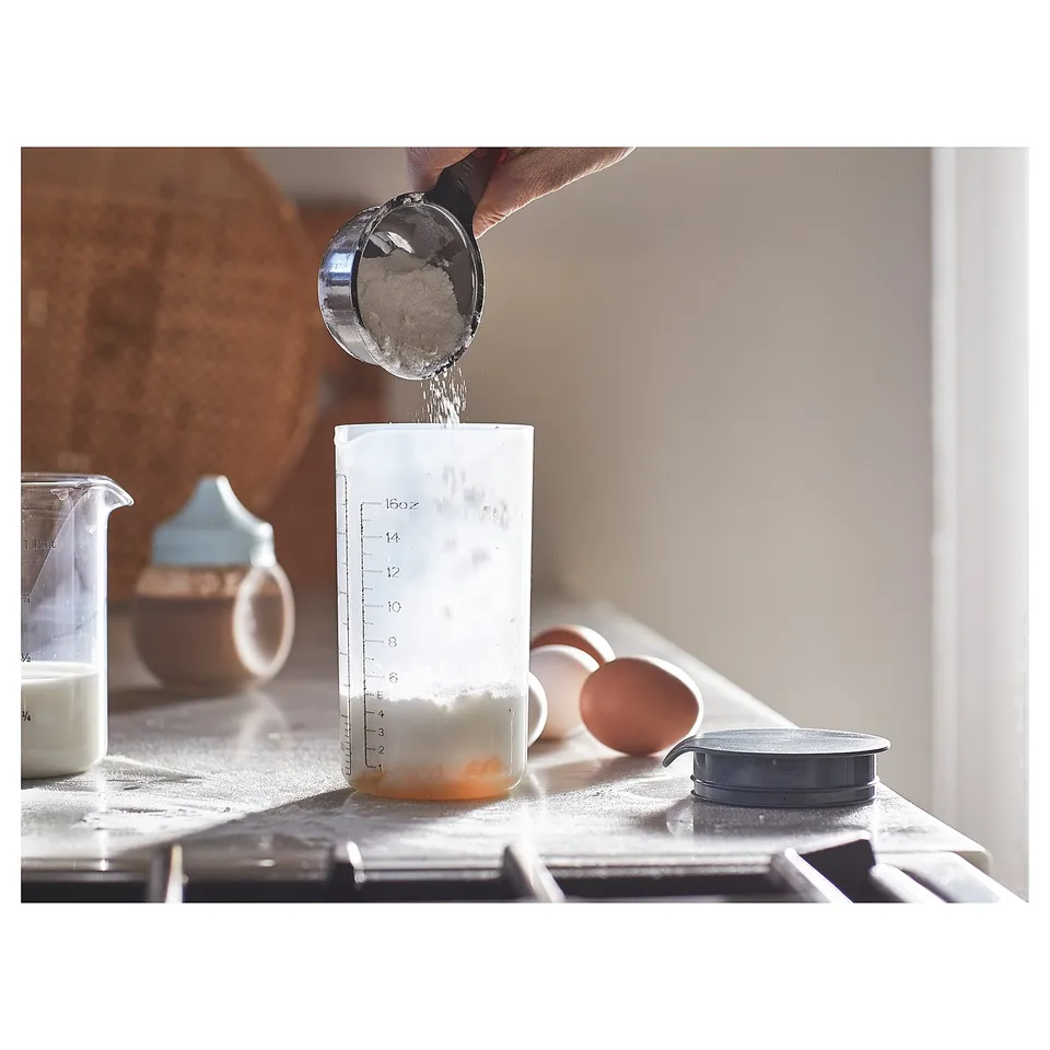 A person shakes powder into a drink or batter using a gradient-marked shaker on a kitchen counter.