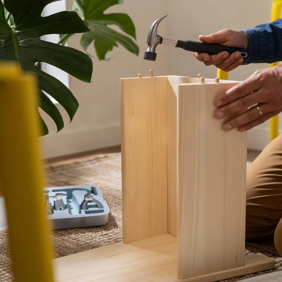 Person using TRIXIG hammer to assemble wooden furniture, showcasing its multipurpose use as a rubber mallet, with tools organised in background.