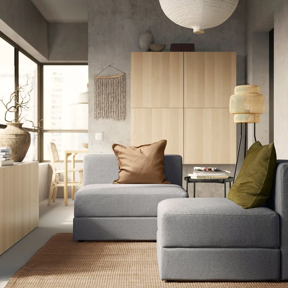 Modern living room with grey JÄTTEBO modular sofa, brown throw pillows, wooden cabinet, and floor lamp. Minimalist decor with natural light.